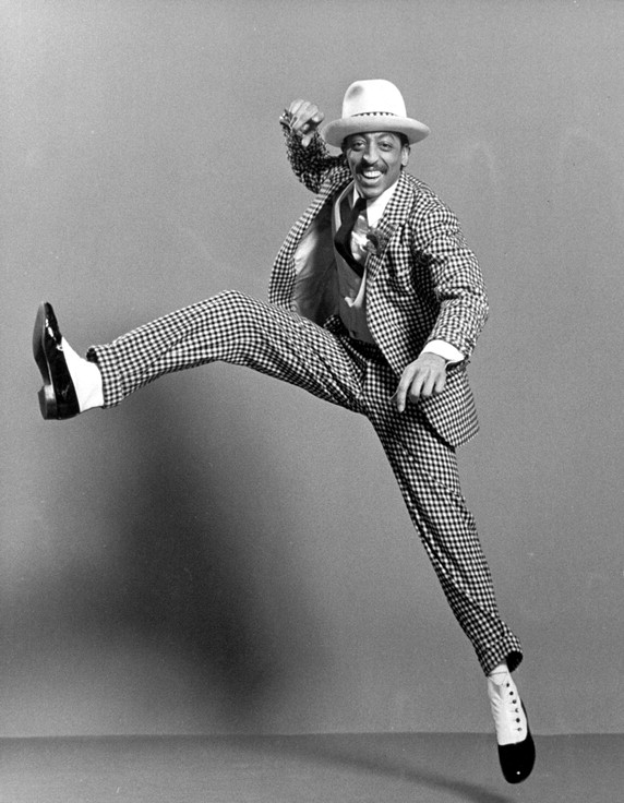 Gregory Hines in a checkered suit, hat, and snazzy tap shoes, kicking up in mid-air.